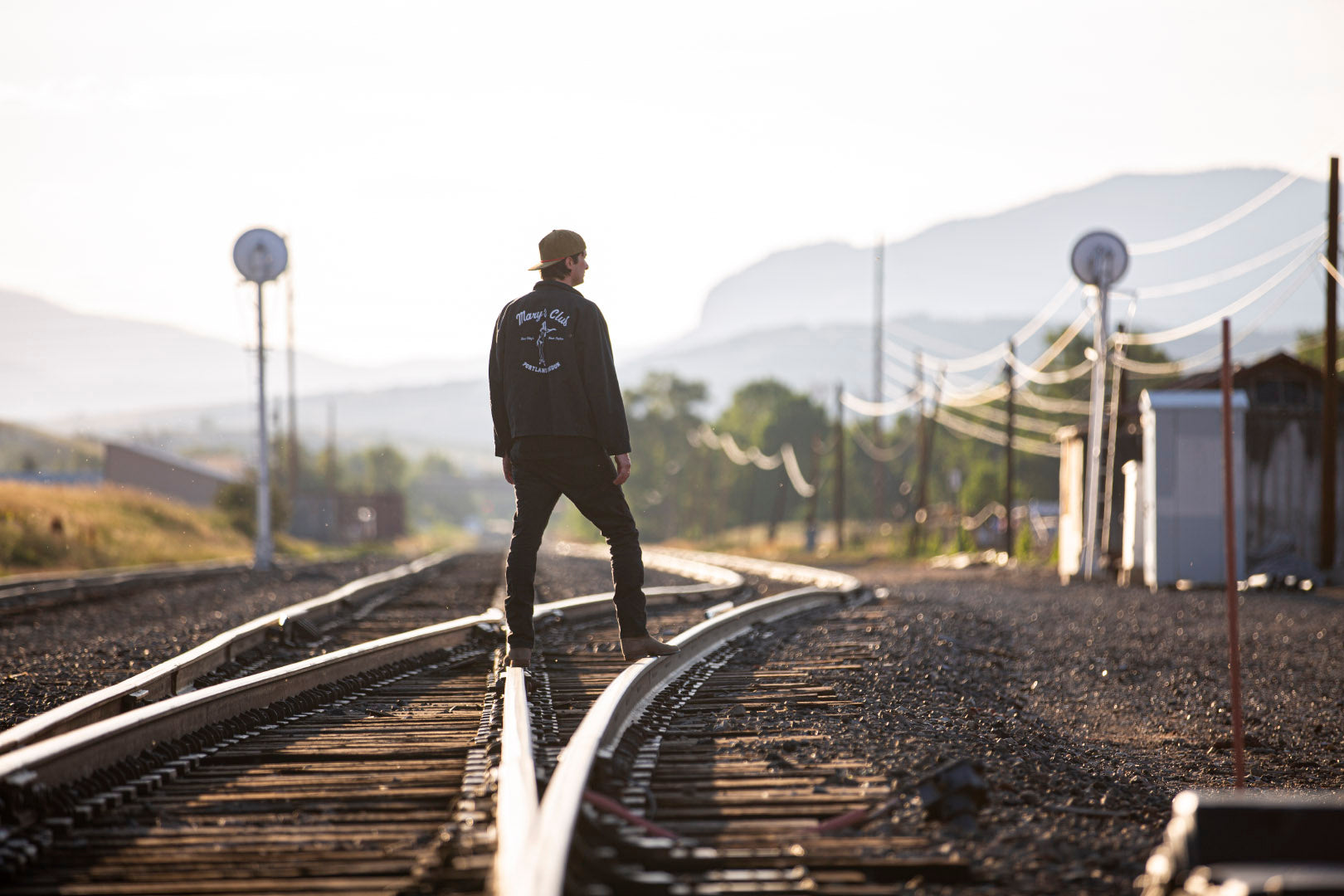 Man standing on train tracks with his back facing the camera in the Blvnk Headwear Gramps Corduroy blank snapback in Crocodile and Red wearing a black Mary's Club gas station jacket.