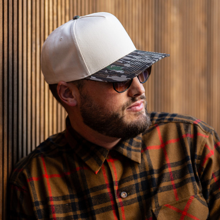 all-groups Blvnk Headwear Origin Vintage Shape blank Snapback in Natural and Realtree Original Camo bill on man in a flannel against a wood slat backdrop.