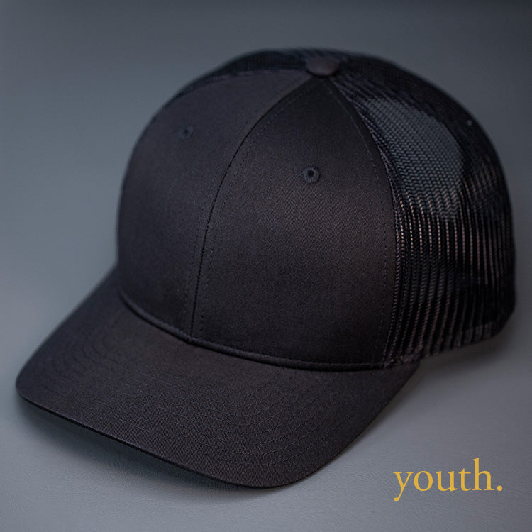 A Youth Sized, Black on Black, Cotton Twill, Mesh Backed, Blank Trucker Hat with a Pre Curved Bill, & Classic Snapback. | Designed by Blvnk Headwear