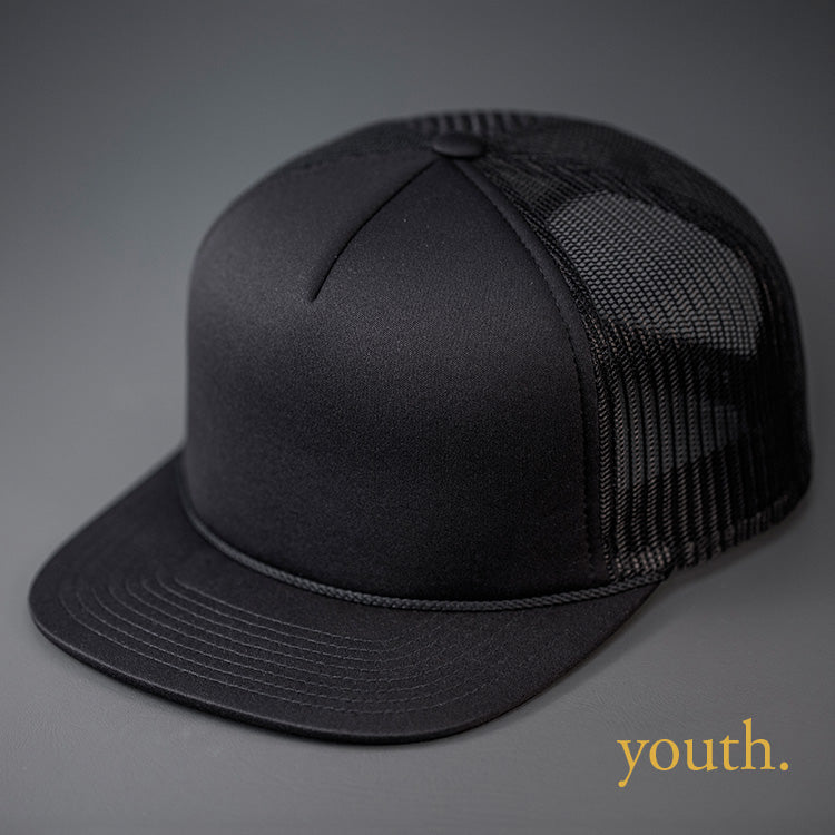 A Youth Sized, Black on Black, Foam Front, Mesh Backed Blank Trucker Hat with a Flat Bill, & Classic Snapback. Designed by Blvnk Headwear.