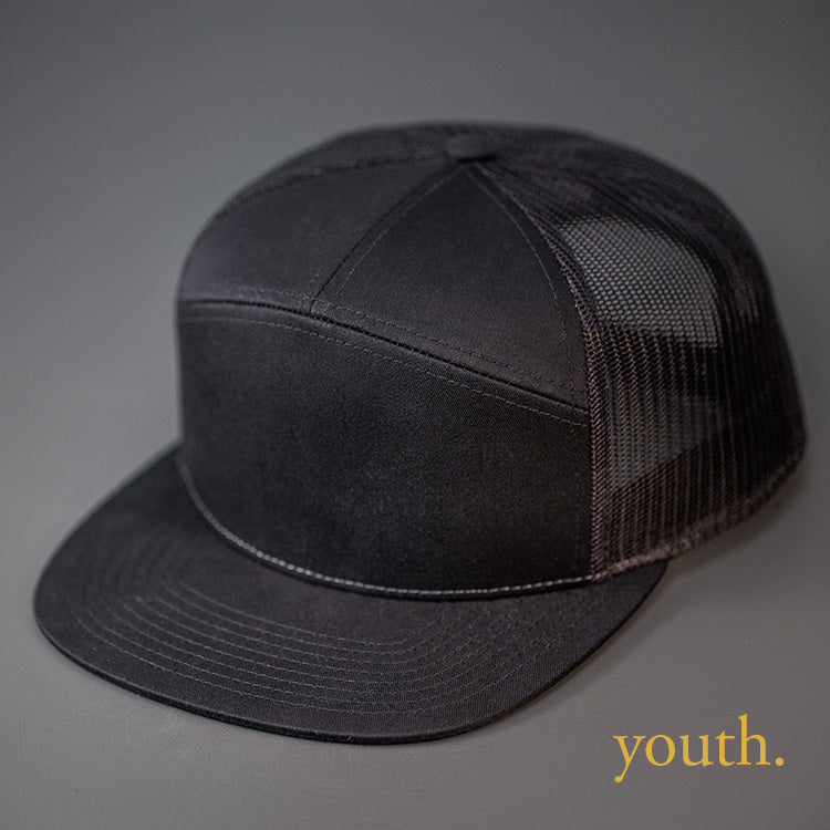 A Youth Sized, Black, Cotton Twill, Mesh Backed Blank 7 Panel Trucker Hat with a Flat Bill, & Classic Snapback. Designed by Blvnk Headwear