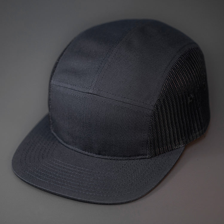 A Black, Cotton Twill/Mesh, Blank 5 Panel Camp Trucker Hat With a Flat Bill, & Woven Nylon Strapback. Designed by Blvnk Headwear.