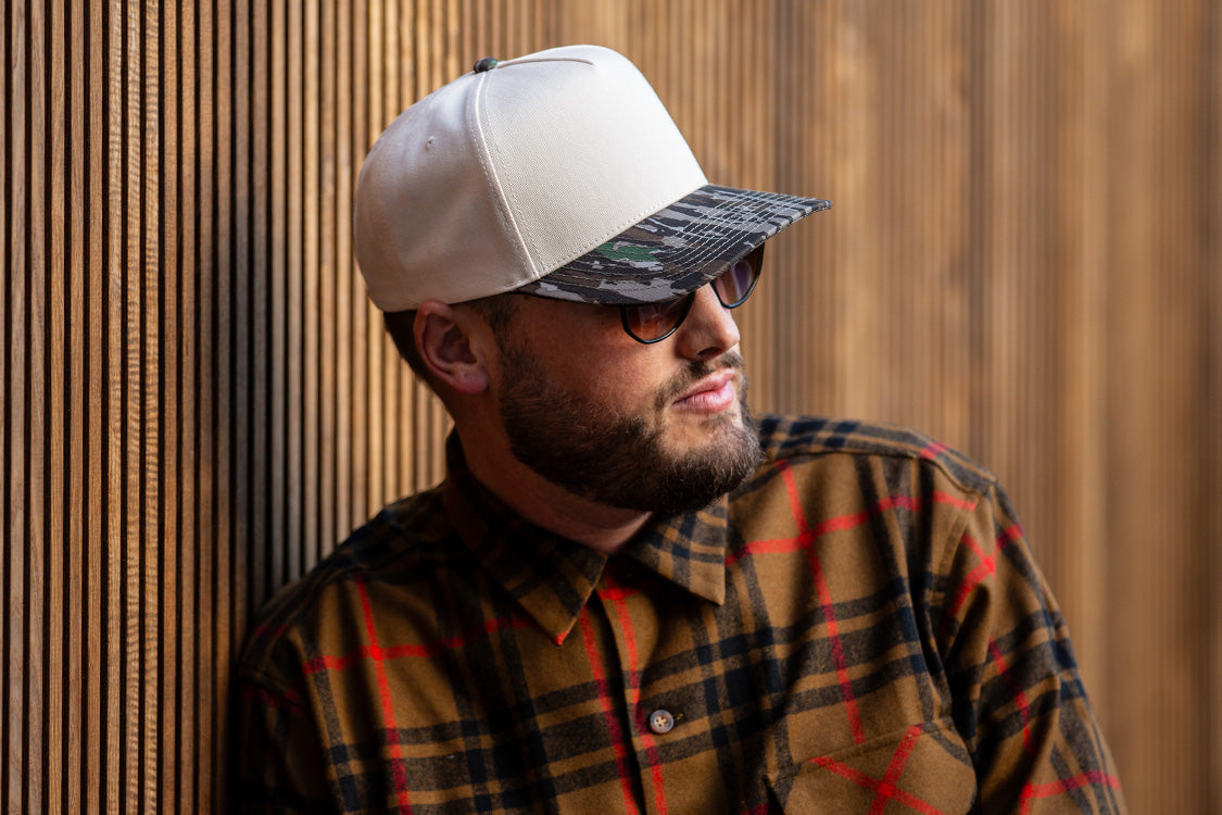 all-groups Blvnk Headwear Origin Vintage Shape blank Snapback in Natural and Realtree Original Camo bill on man in a flannel against a wood slat backdrop.