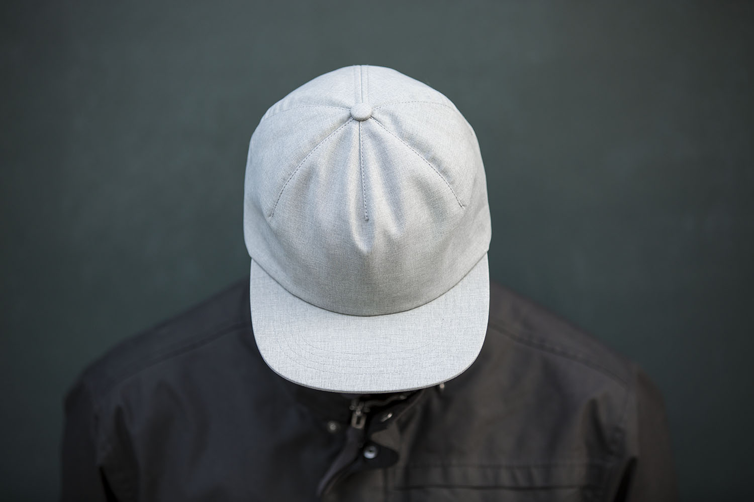 A man wearing Blvnk Headwear's The One Panel blank strap back hat in Heather Grey standing against a dark green wall in a black jacket.