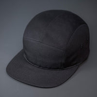 A Black, Cotton Twill, Blank 5 Panel Camp Hat With a Flat Bill, & Woven Nylon Strapback.  Designed by Blvnk Headwear