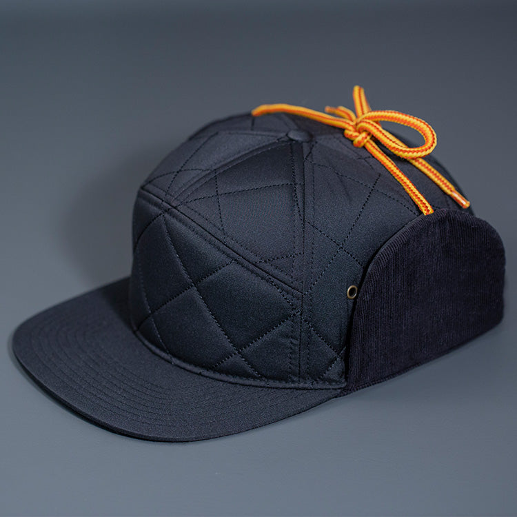 A Satin Lined, Black, Performance Diamond Quilted Blank 7 Panel Hat W/ Navy Corduroy Earflaps & Vegan Leather Strapback W/ Brass Clasp.
