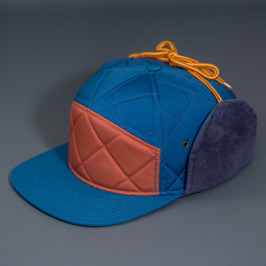 A Satin Lined, Old Blue & Rust, Performance Diamond Quilted Blank 7 Panel Hat W/ Navy Corduroy Earflaps & Vegan Leather Strapback W/ Brass Clasp.