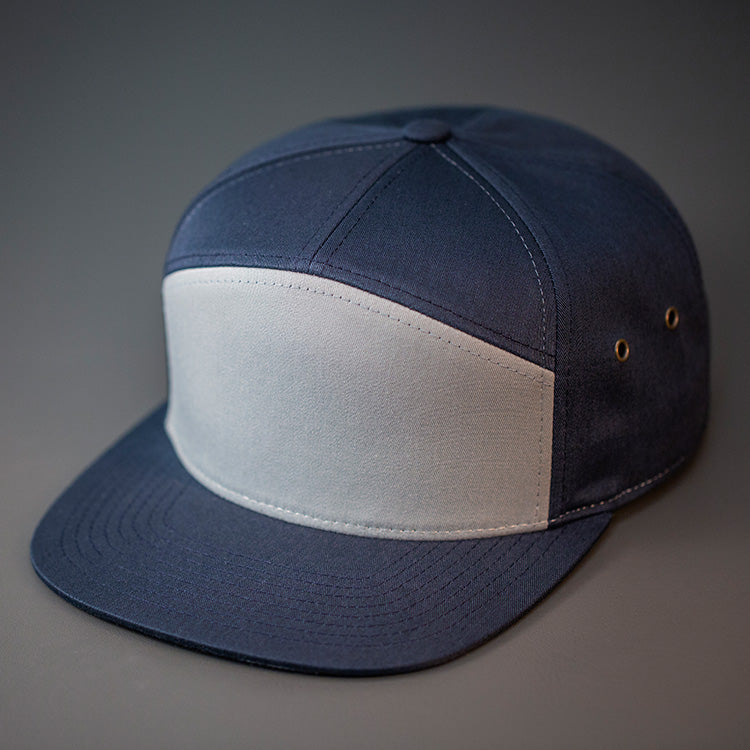 A Pigeon Grey & Navy Blue colored, Cotton Twill, Blank 7 Panel Hat with a Flat Bill, Brass Details, & Vegan Leather Strapback.  Designed by Blvnk Headwear