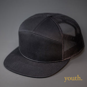 A Youth Sized, Black, Cotton Twill, Mesh Backed Blank 7 Panel Trucker Hat with a Flat Bill, & Classic Snapback.  Designed by Blvnk Headwear